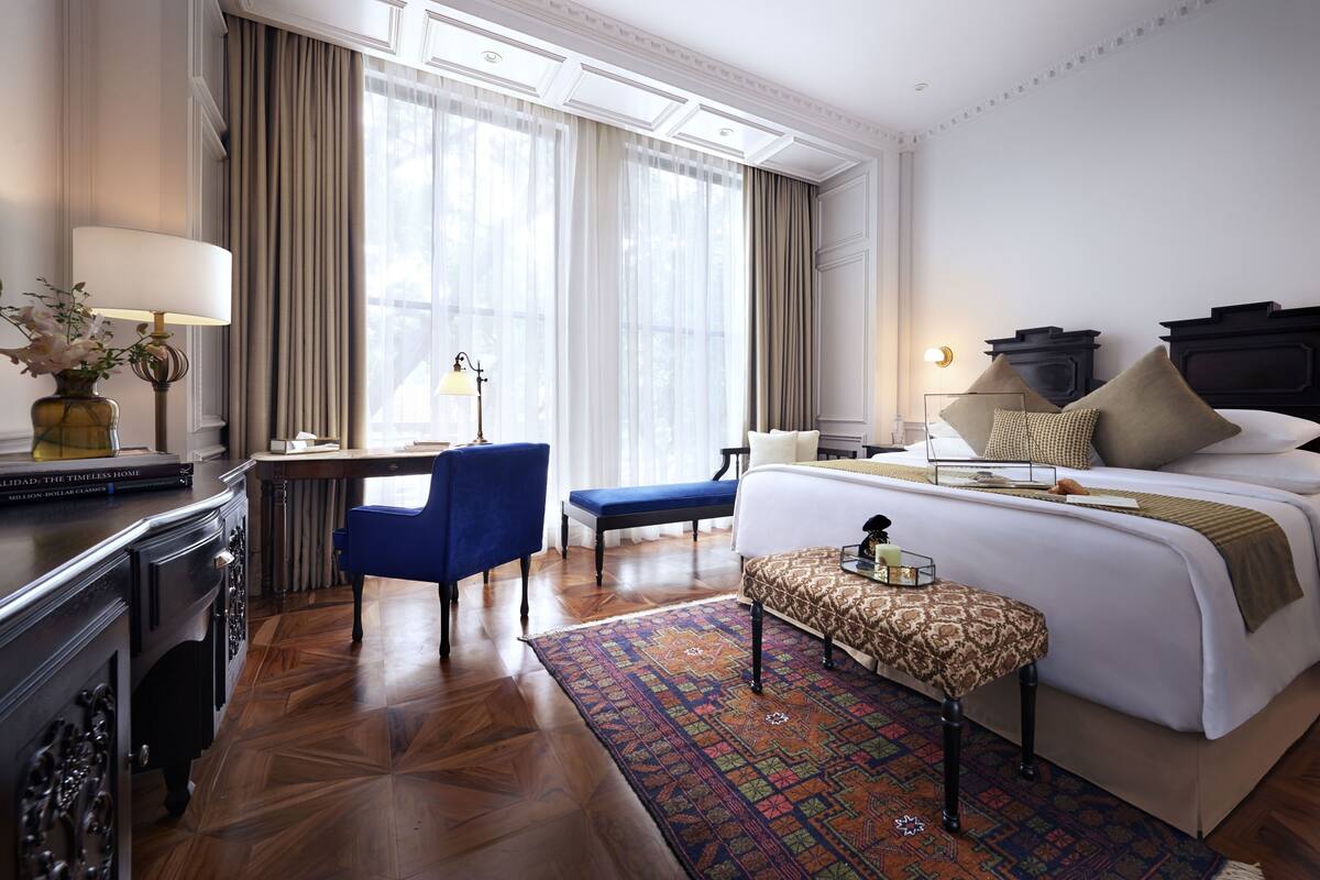 A hotel room with a bed, a desk with a chair, a small table with a lamp, and a window with curtains.
