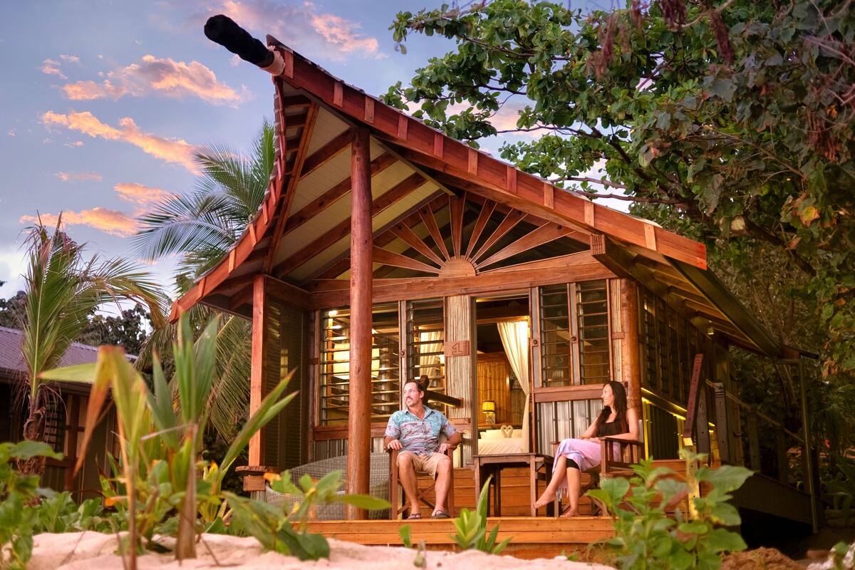 A wooden structure with a covered porch, a bed, and a window with curtains.