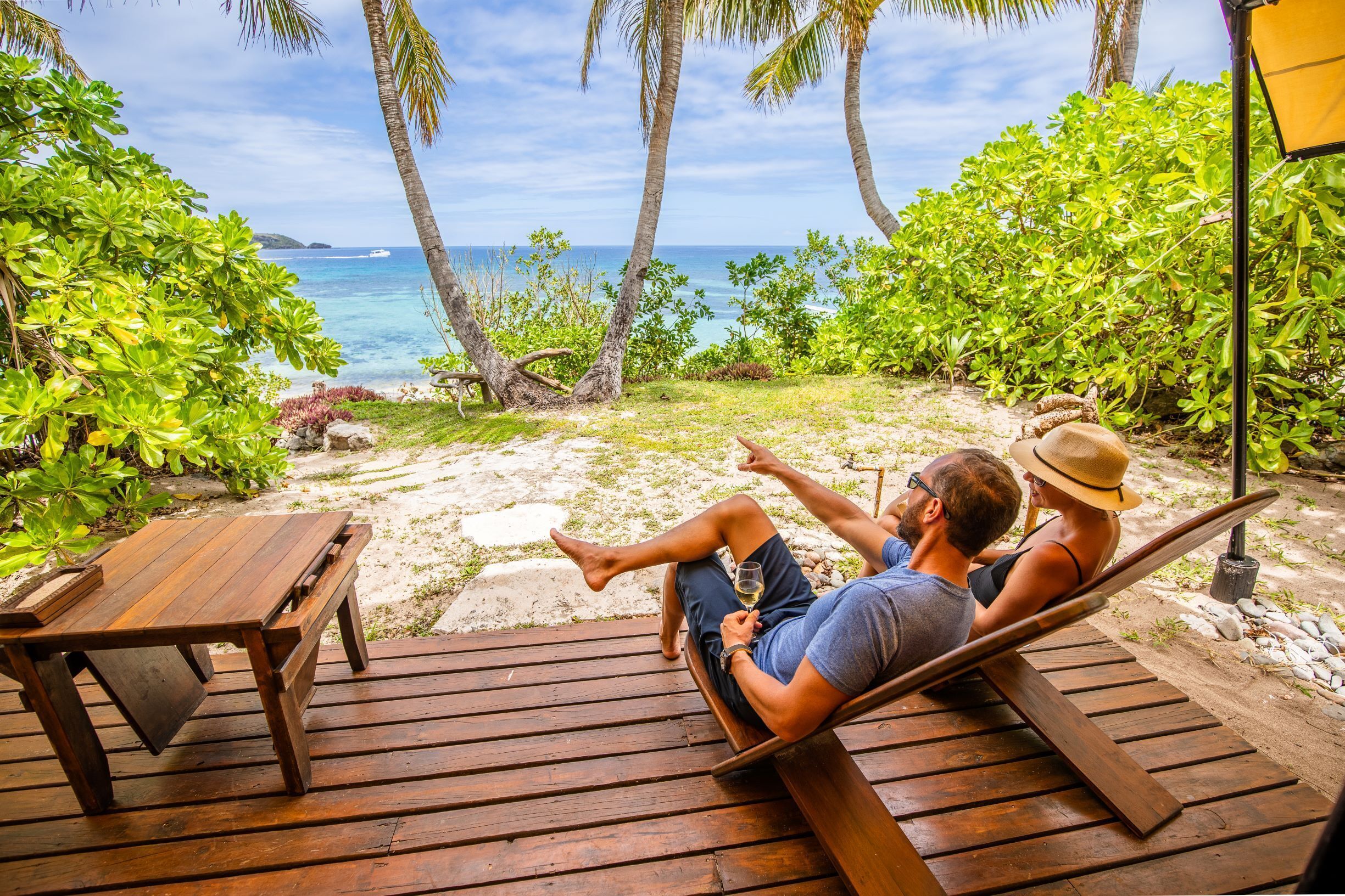 A wooden deck with a hammock, a wooden table, and a view of the ocean and palm trees.
