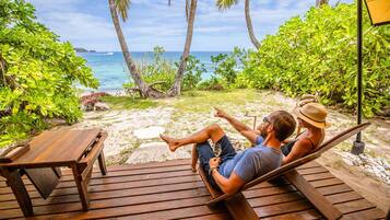 A wooden deck with a hammock, a wooden table, and a view of the ocean and palm trees.