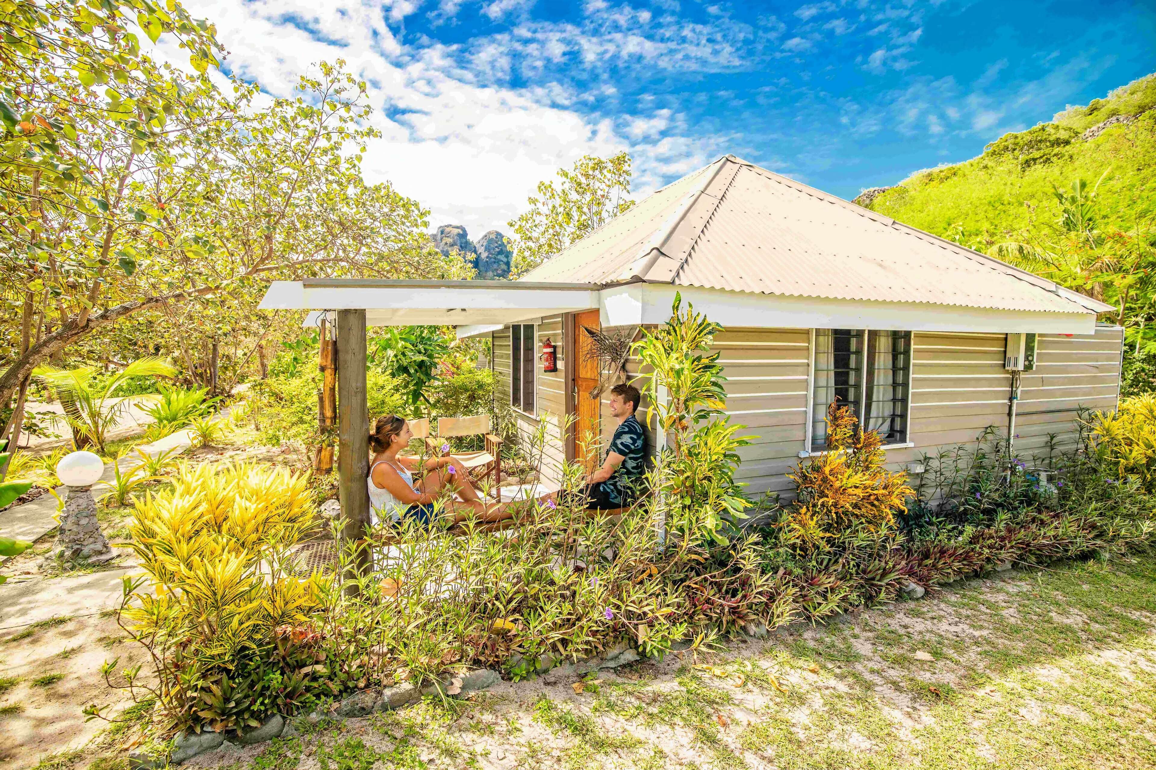 A small, single-story house with a porch, surrounded by lush greenery and a clear sky.