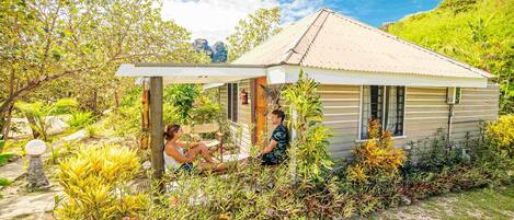 A small, single-story house with a porch, surrounded by lush greenery and a clear sky.