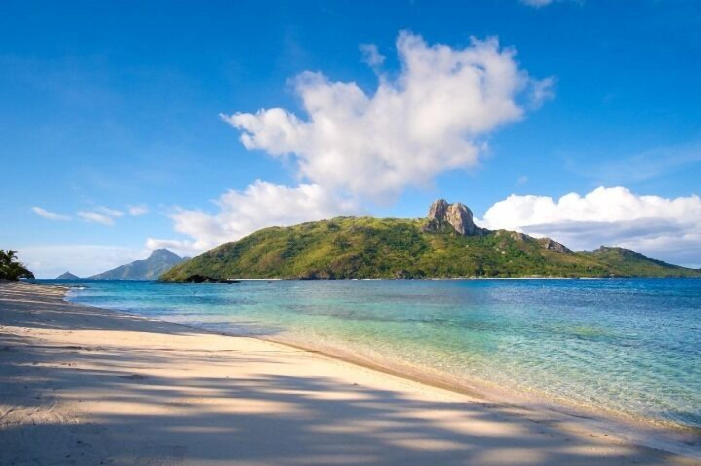 A beach with clear turquoise water, a sandy shore, and a green hill with a rocky peak in the background.