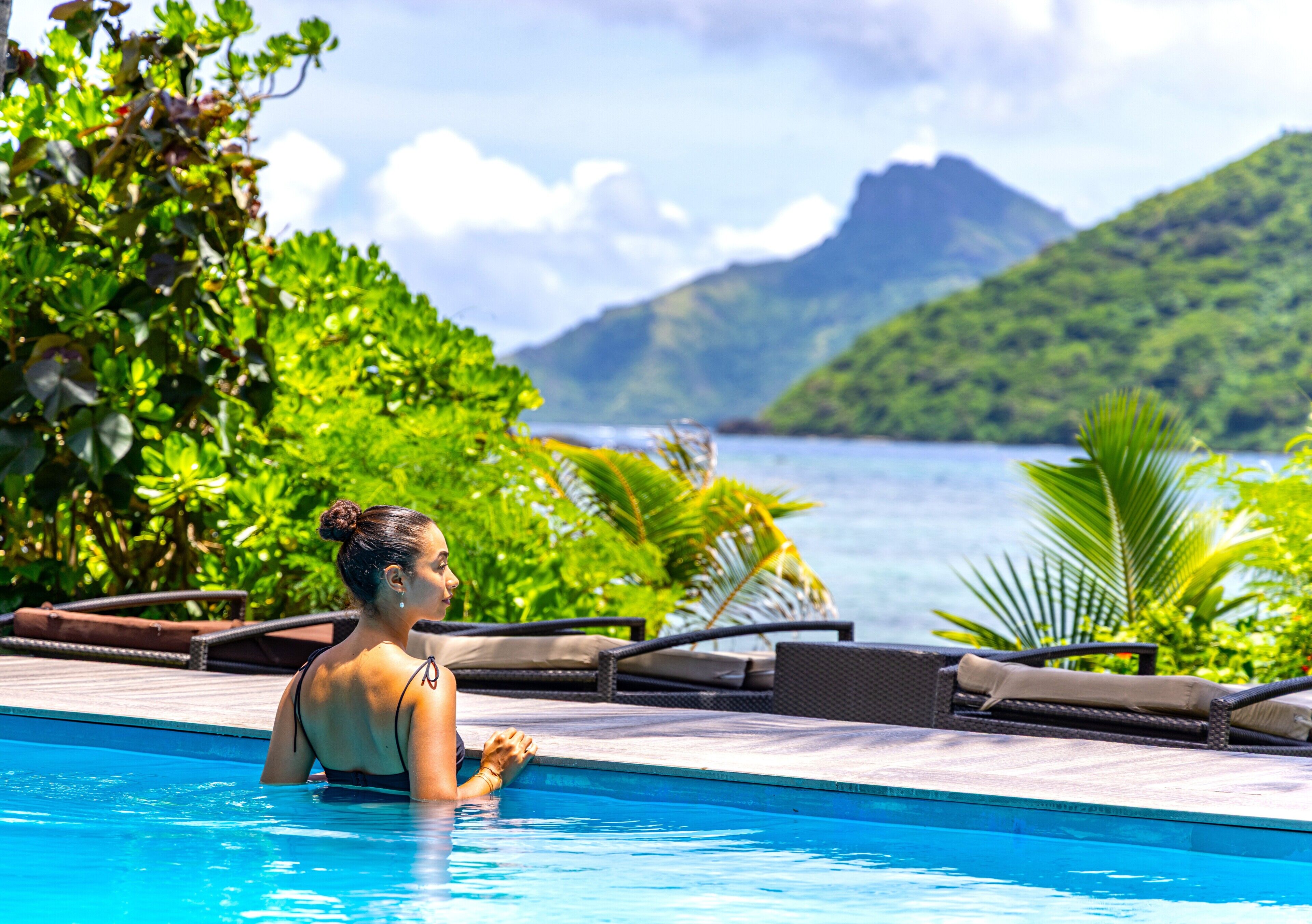 A poolside area with lounge chairs, a pool, and a view of a mountain and the sea.