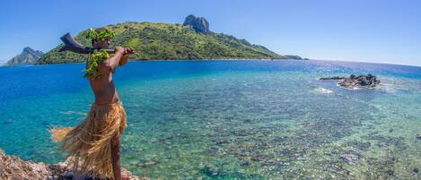 A person standing on a rocky outcrop by a clear blue sea with a mountainous backdrop.