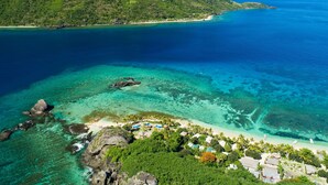 Une île tropicale aux eaux d’un bleu cristallin, avec une plage rocheuse et une petite zone touristique comprenant plusieurs bâtiments et des palmiers.