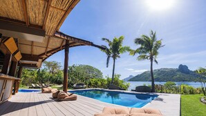 Un espace piscine avec des chaises longues, une structure en bambou et une vue sur la mer et les montagnes.