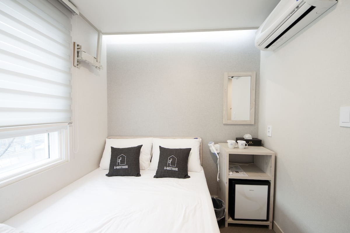 A hotel room with a bed, two pillows with 'K-GUESTHOUSE' logo, a bedside table with a cup and a small fridge, and a window with blinds.