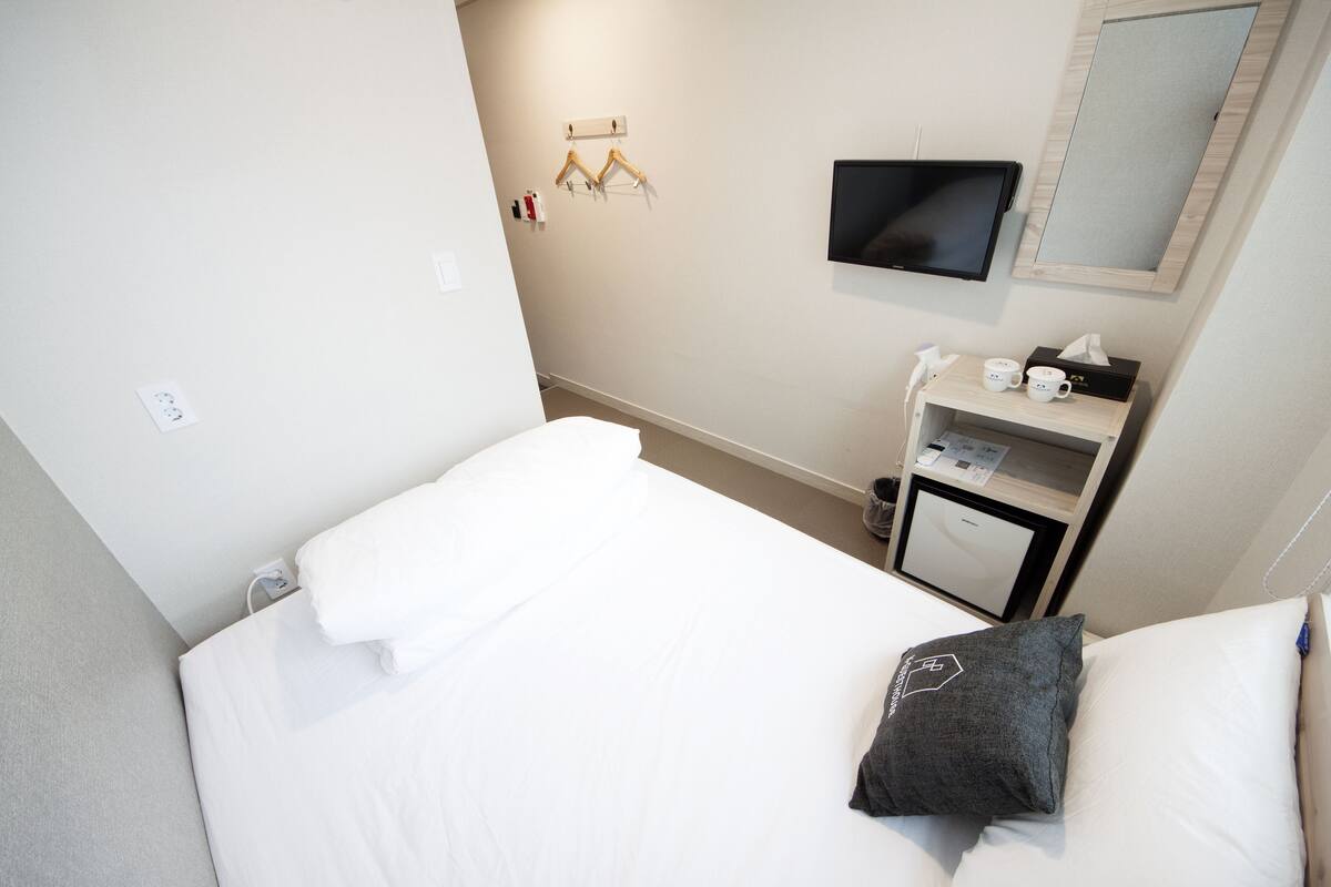 A hotel room with a bed, a small desk with a mirror, a TV, and a wall-mounted shelf with cups and a tissue box.