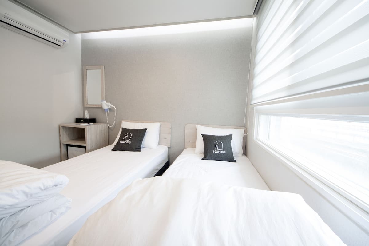 A hotel room with two single beds, a bedside table with a lamp, a mirror, and a window with blinds.