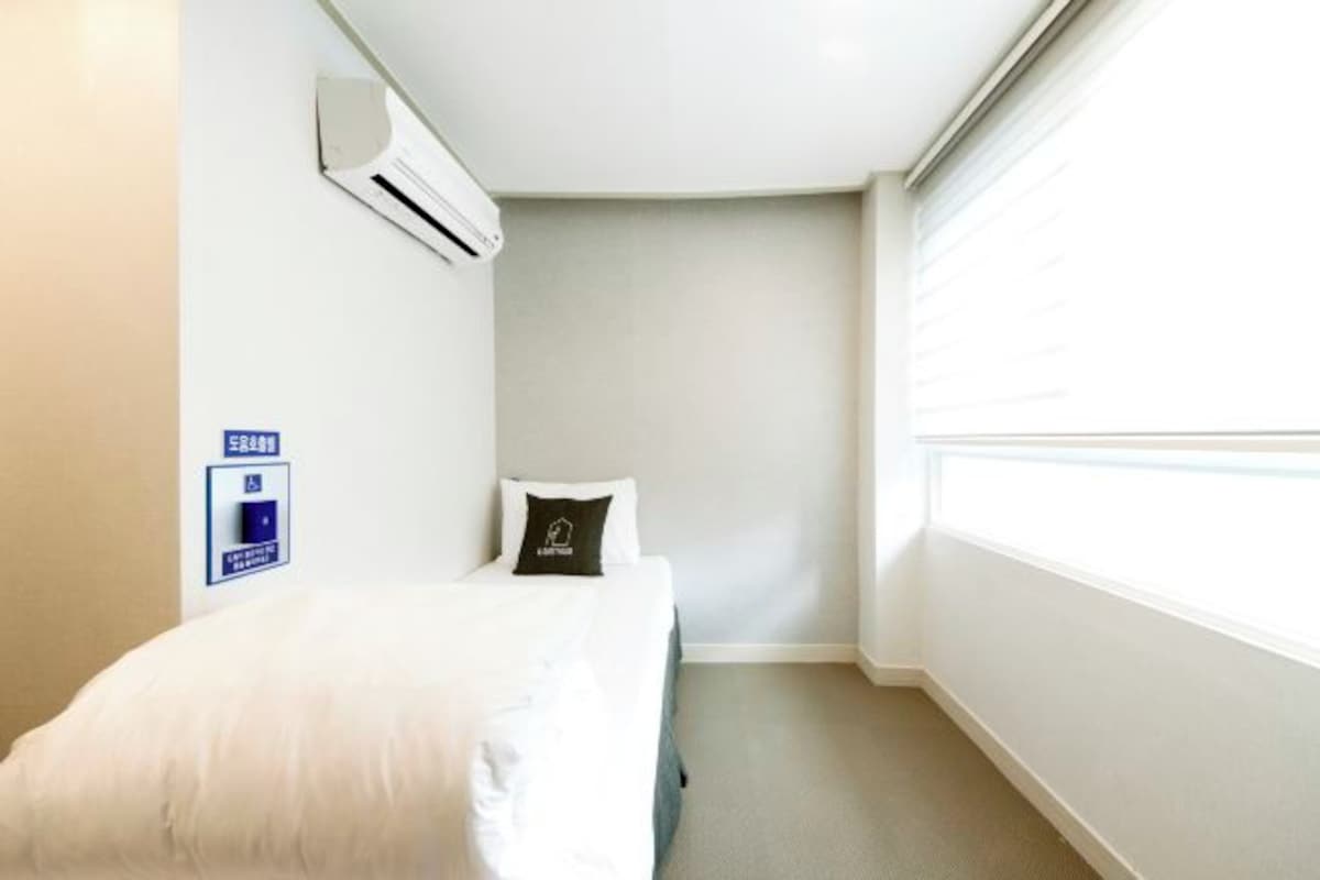 A small hotel room with a single bed, a window, and an air conditioning unit on the wall.
