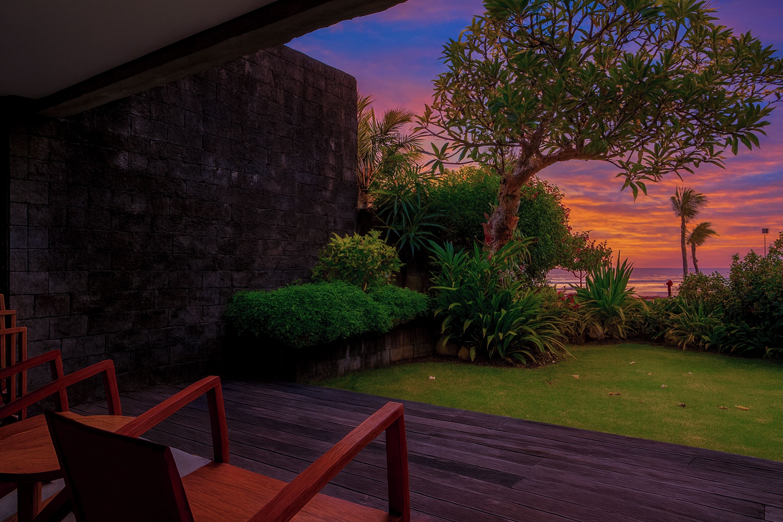 A patio with wooden furniture and a view of a sunset over the ocean.