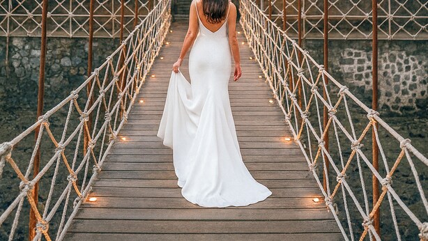 A wooden bridge with rope railings and a hanging lantern, leading to a building with a stone wall and greenery.