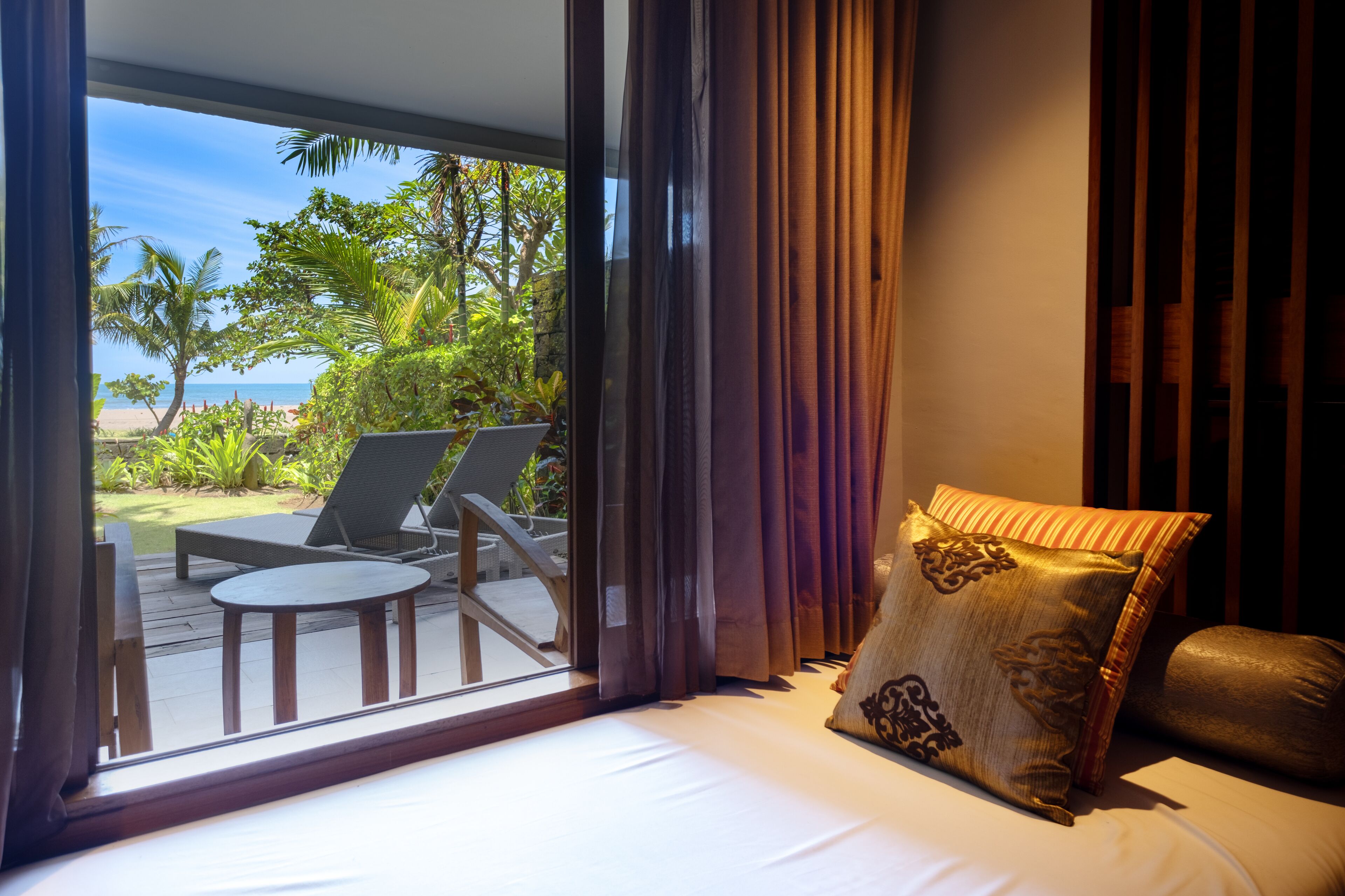 A bedroom with a large window, a bed, and a view of a deck with lounge chairs and a table.