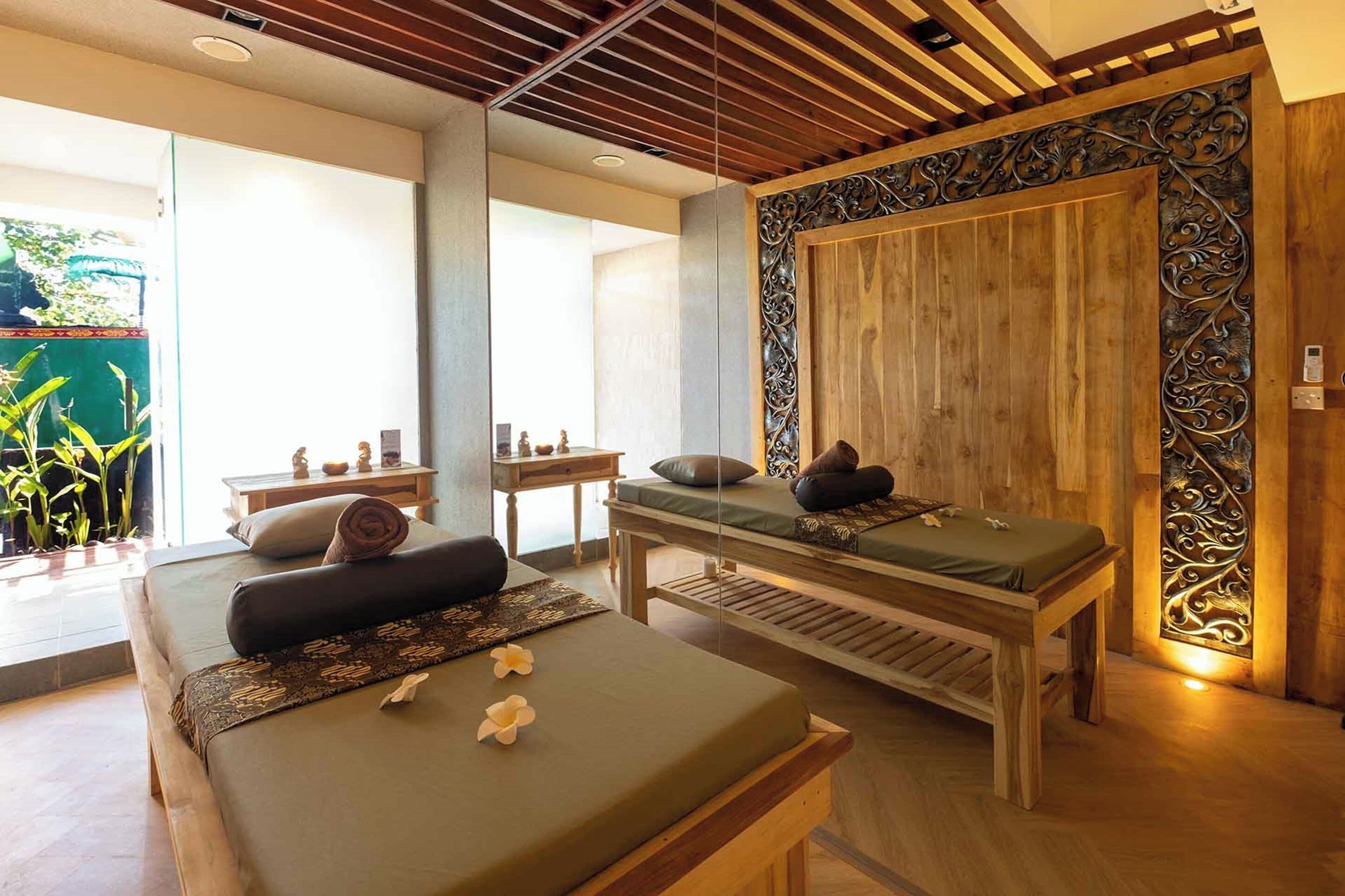 A spa room with two massage tables, a wooden wall with decorative paneling, and a view of greenery outside.