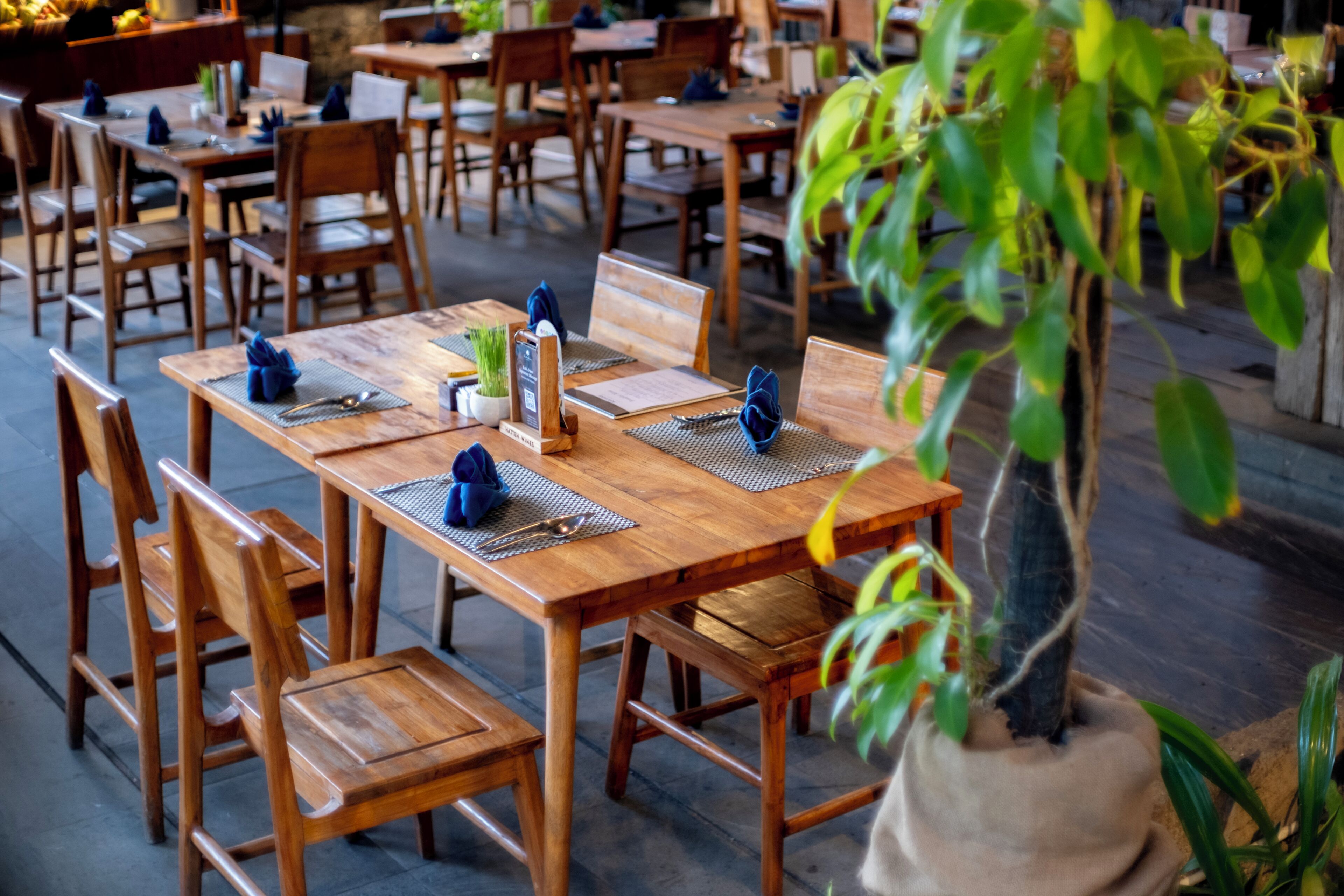 A restaurant with wooden tables and chairs, some with blue napkins and cutlery.