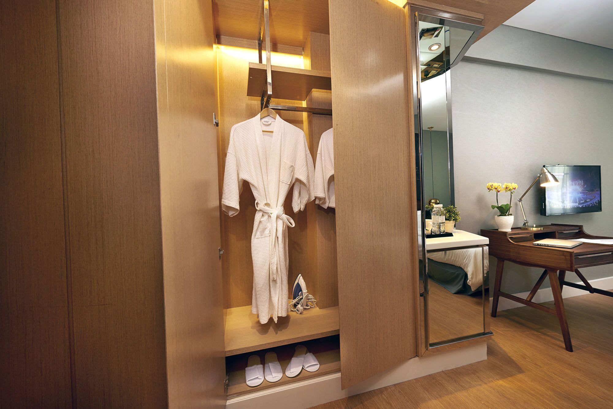 A walk-in closet with hanging robes and slippers, a desk with a lamp, and a TV on the wall.