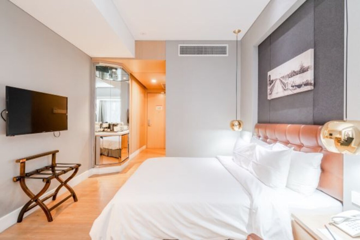 A modern hotel room with a large bed, a flat-screen TV mounted on the wall, a bedside table with a lamp, and a view of the bathroom through an open door.
