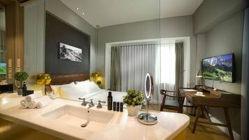 A modern hotel room with a large bathroom sink, a bed with a headboard, a desk with a chair, a television, and a window with curtains.