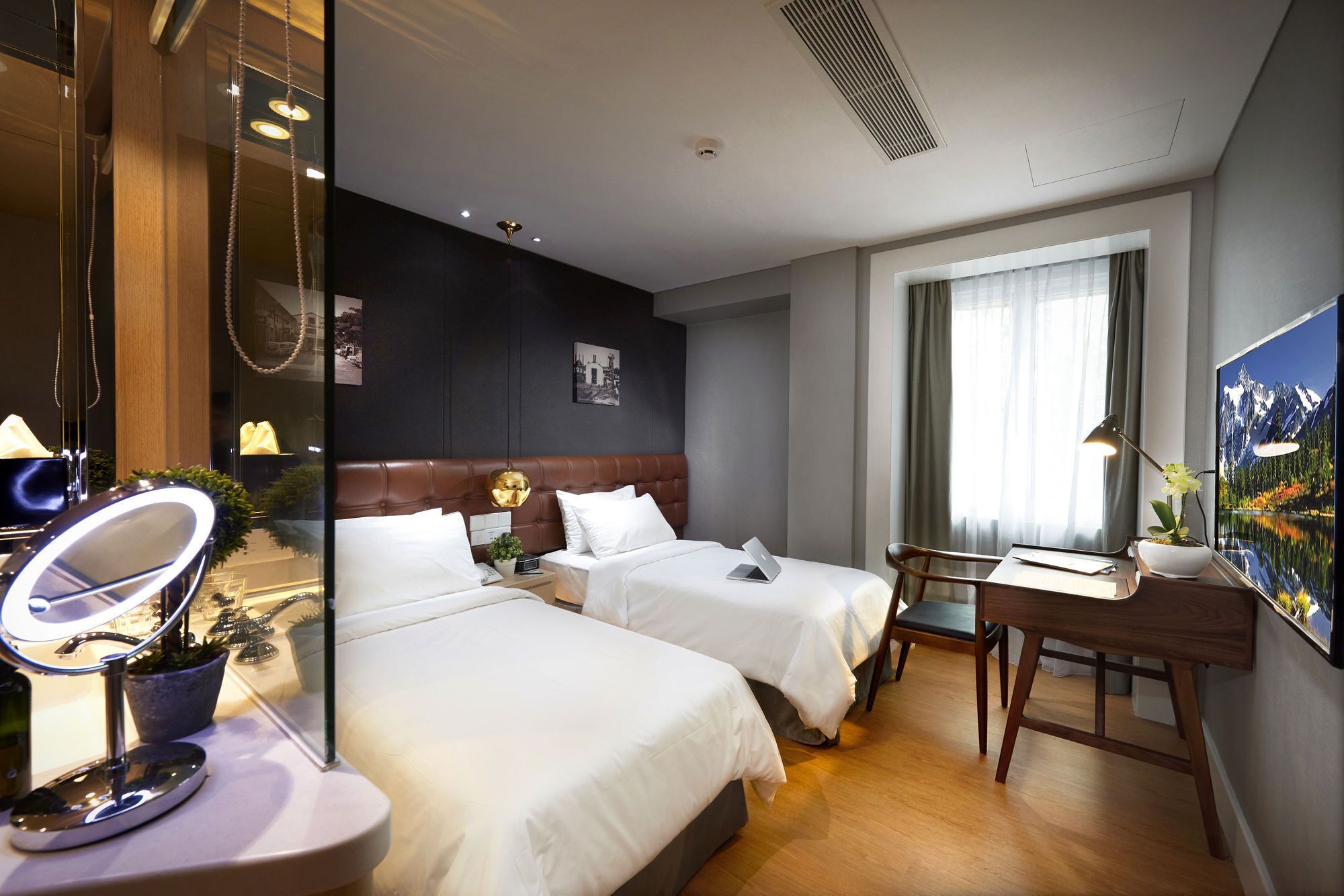 A hotel room with two beds, a desk with a chair, a large mirror, and a painting on the wall.