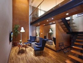Un interior moderno con piso de madera, sillones azules, una pequeña mesa con lámpara y una escalera.