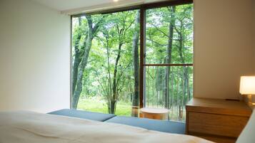 A bedroom with a large window offering a view of a forest. There is a bed with a blue headboard, a wooden nightstand, and a lamp.