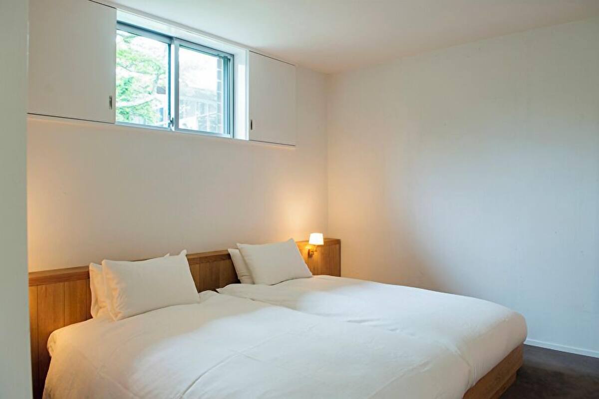 A single bed with a wooden headboard, a window above it, and a wall-mounted light.