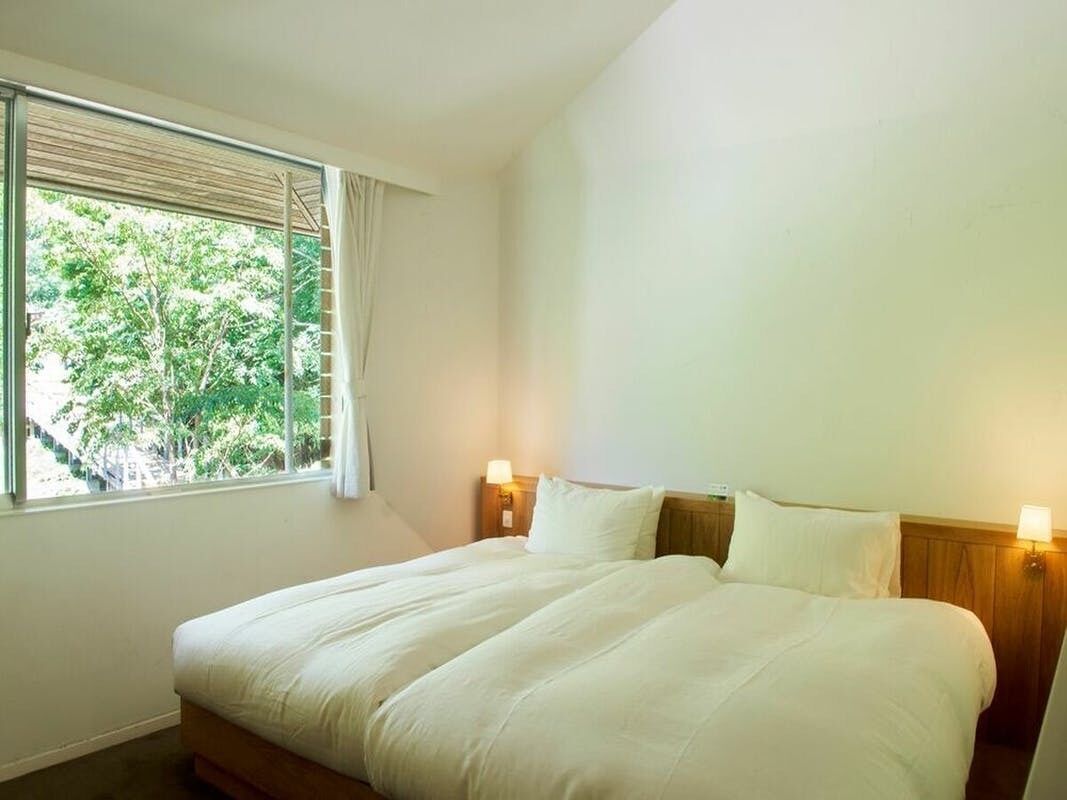 A hotel room with a large bed, two bedside lamps, a window with blinds, and a view of trees.