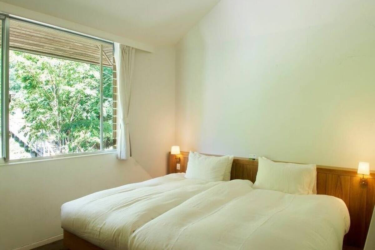 A hotel room with a large bed, two bedside lamps, a window with blinds, and a view of trees.