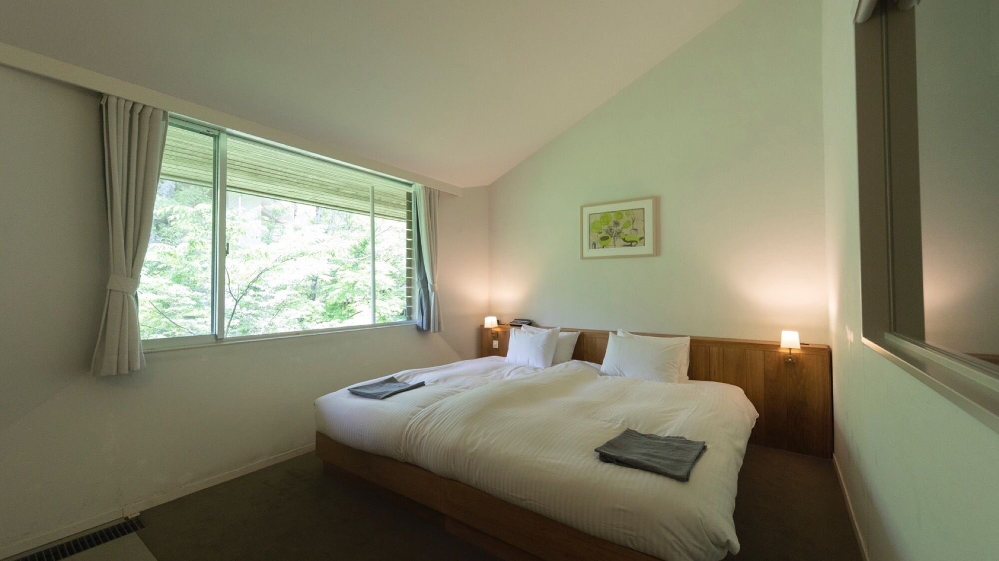 A hotel room with a large bed, a window with curtains, a nightstand with a lamp, and a framed picture on the wall.