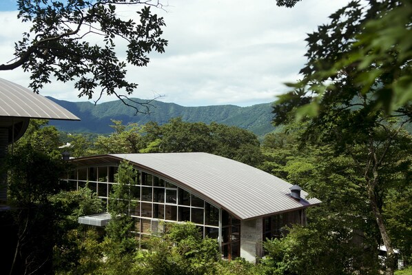 A modern building with a sloped roof surrounded by dense forest and mountains in the background. - Hakone Retreat Före (Hakone)