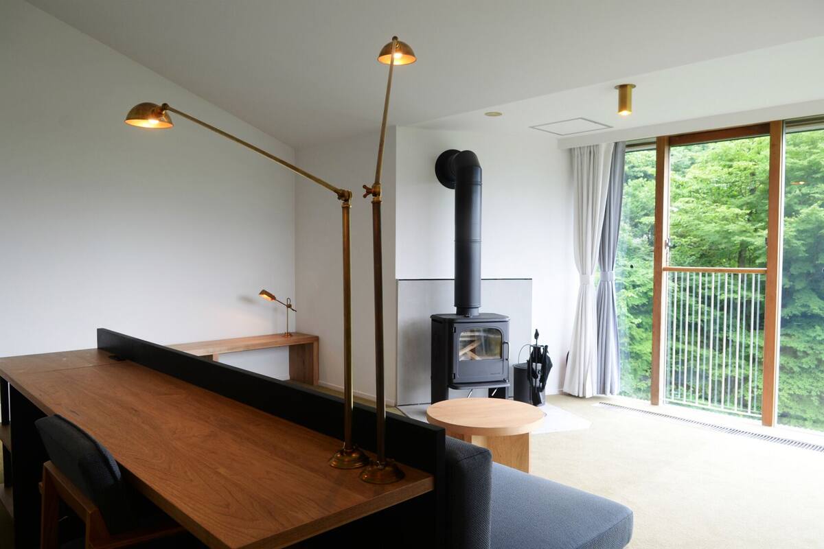 A modern living room with a wooden table, two floor lamps, a black wood-burning stove, and a large sliding glass door leading to a balcony with a forest view.