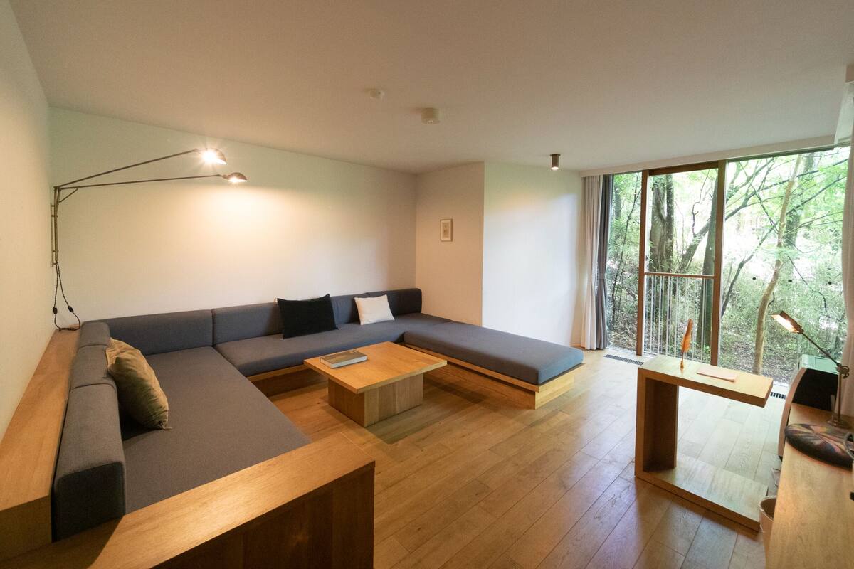 A modern living room with a sectional sofa, a wooden coffee table, and a large window offering a view of trees.