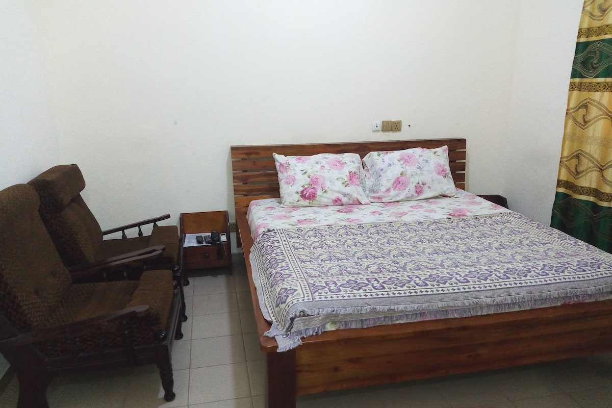 A single bed with a floral-patterned bedspread and two pillows, a wooden nightstand with a lamp, and a brown armchair.