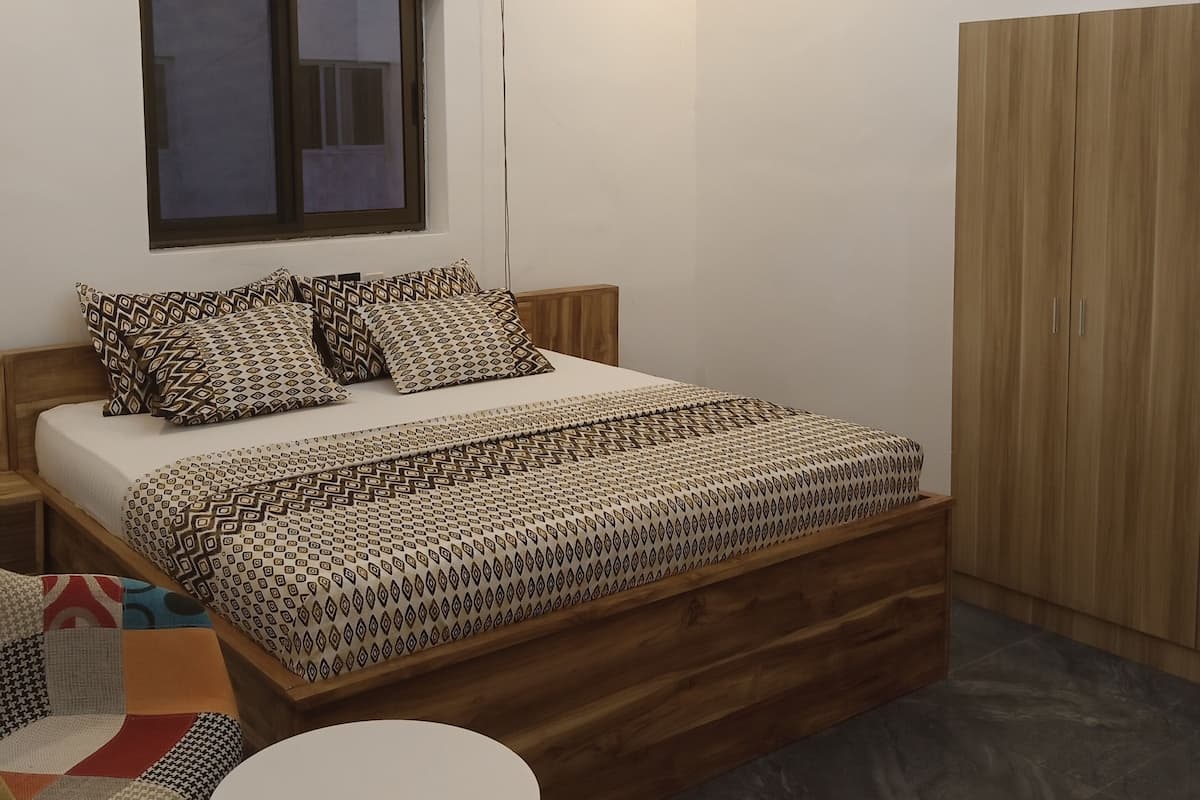 A bedroom with a bed, a window, an air conditioner, a wardrobe, a round table, and a chair.