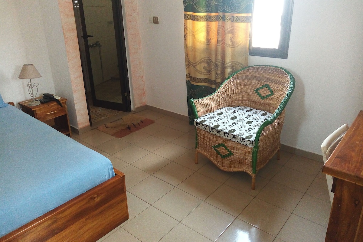 A room with a bed, a chair, a small table, a lamp, and a door leading to a bathroom.