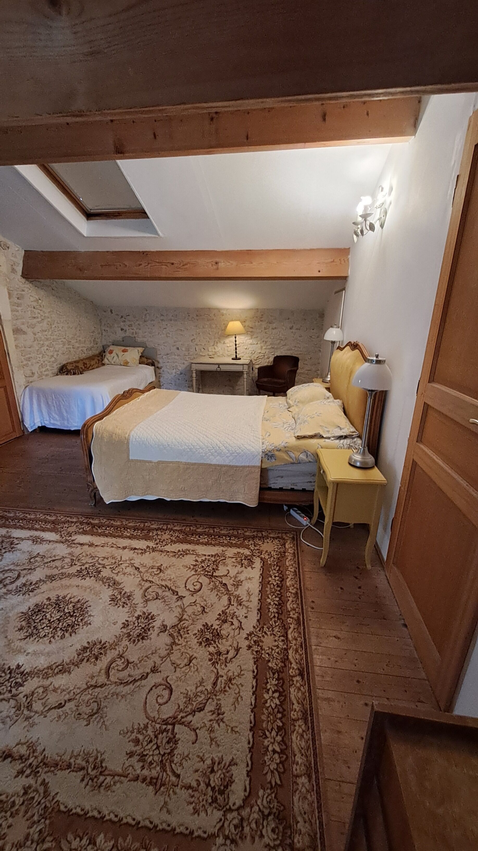 Un dormitorio con dos camas, techo de madera, pared de piedra, una alfombra y una mesita de noche con una lámpara.