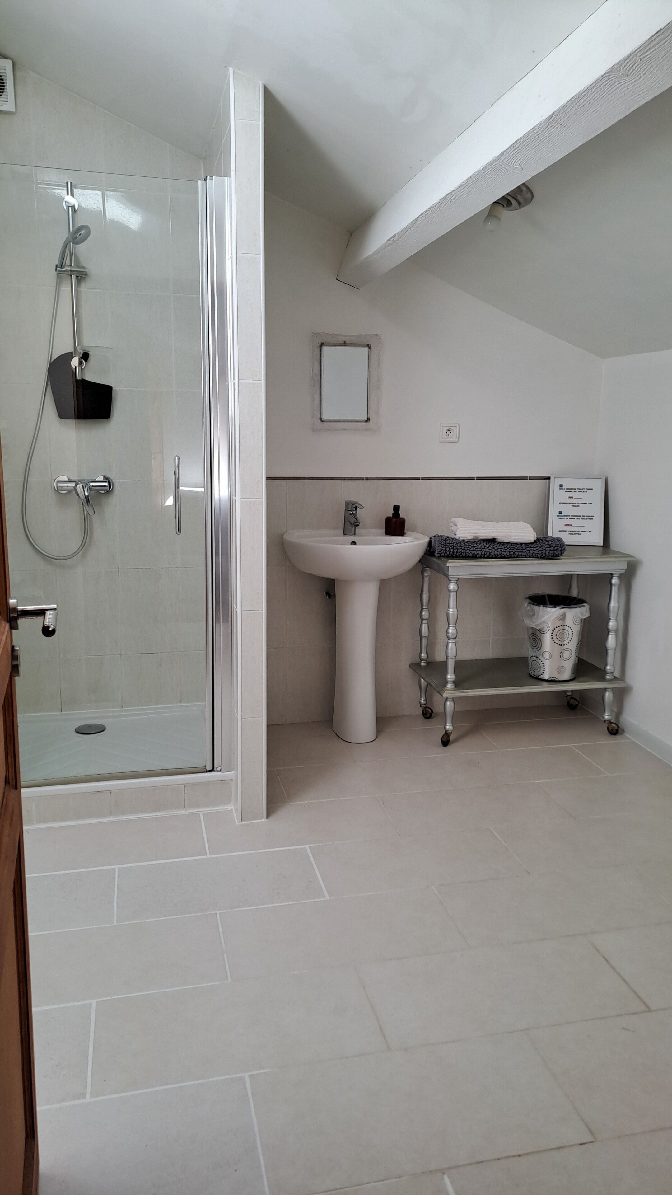 A modern bathroom with a glass shower enclosure, a pedestal sink, and a small vanity table with a trash bin.