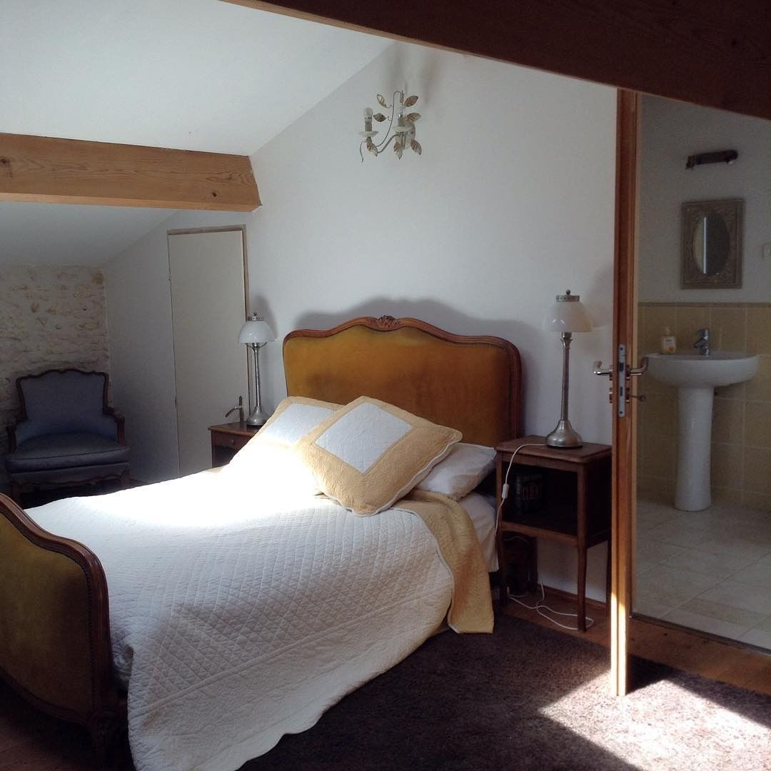 A bedroom with a wooden headboard, a bed with white and beige bedding, two bedside lamps, a chair, and a bathroom visible through an open door.