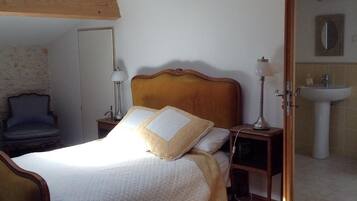 A bedroom with a wooden headboard, a bed with white and beige bedding, two bedside lamps, a chair, and a bathroom visible through an open door.