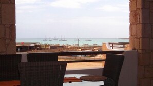 Un espace extérieur aménagé, abrité et doté de chaises en osier et d’une table, offrant une vue sur un port de plaisance avec ses bateaux et ses bâtiments.
