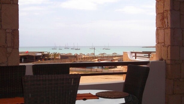 Un espace extérieur aménagé, abrité et doté de chaises en osier et d’une table, offrant une vue sur un port de plaisance avec ses bateaux et ses bâtiments.