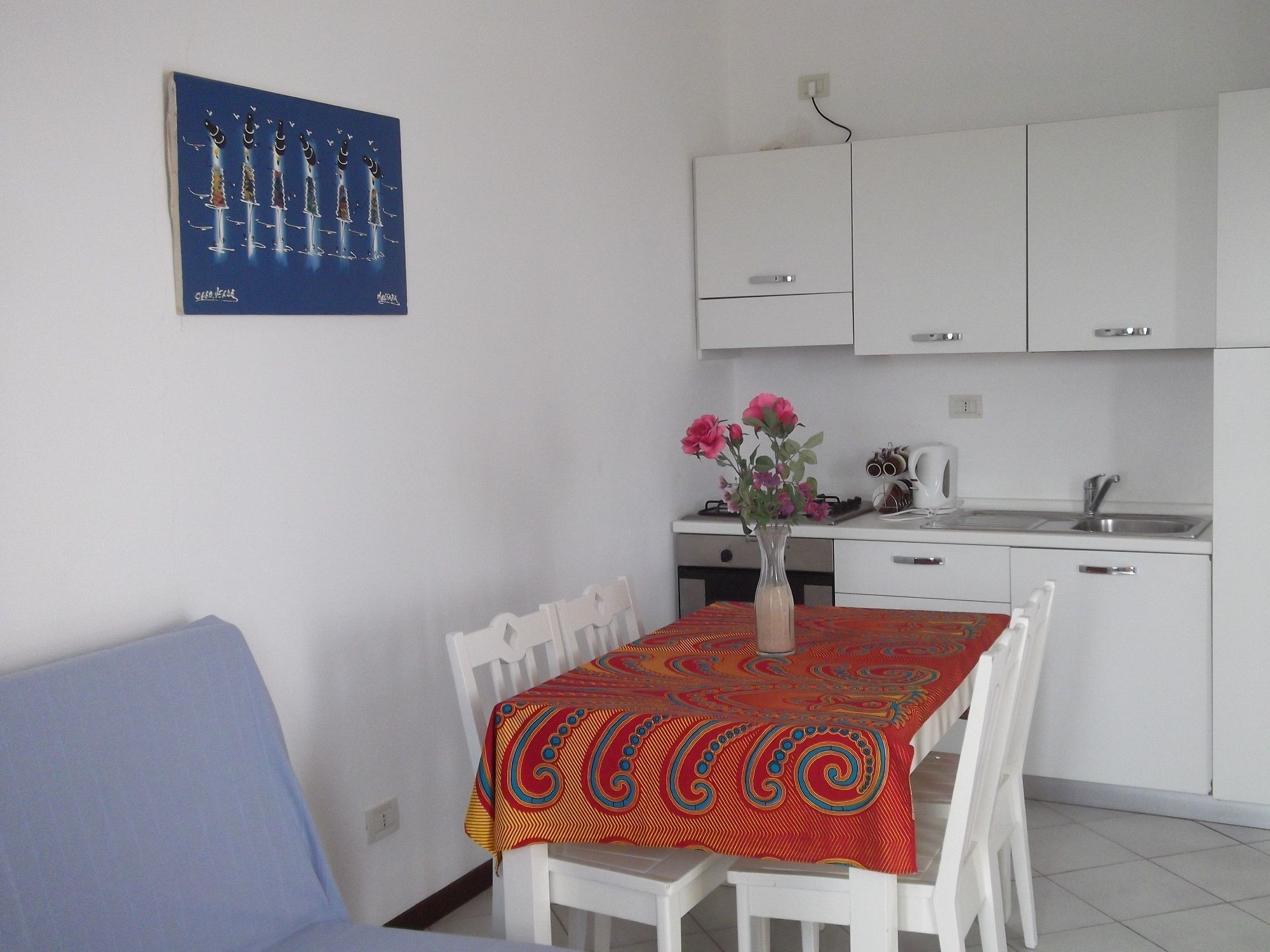 A small, clean kitchen with white cabinets, a dining table with a colorful tablecloth, and a vase with flowers on the table.
