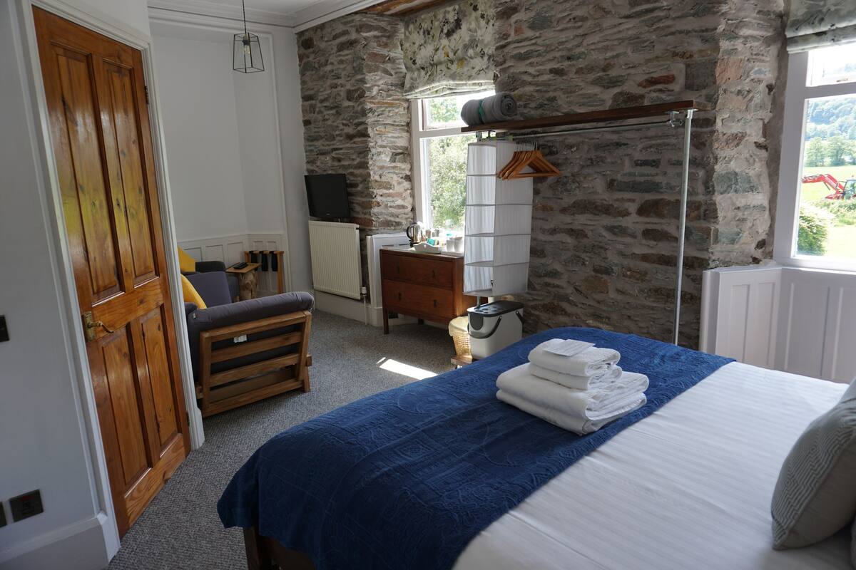 A bedroom with a stone wall, a wooden wardrobe, a bedside table with a lamp, and a window with a view of the outside.