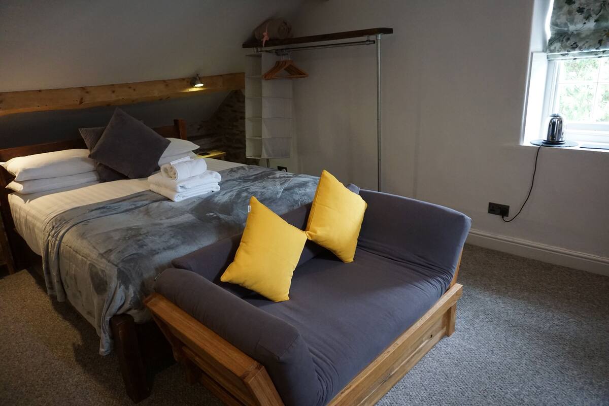 A bedroom with a bed, a sofa, a wooden headboard, a window with a view, and a hanging clothes rack.