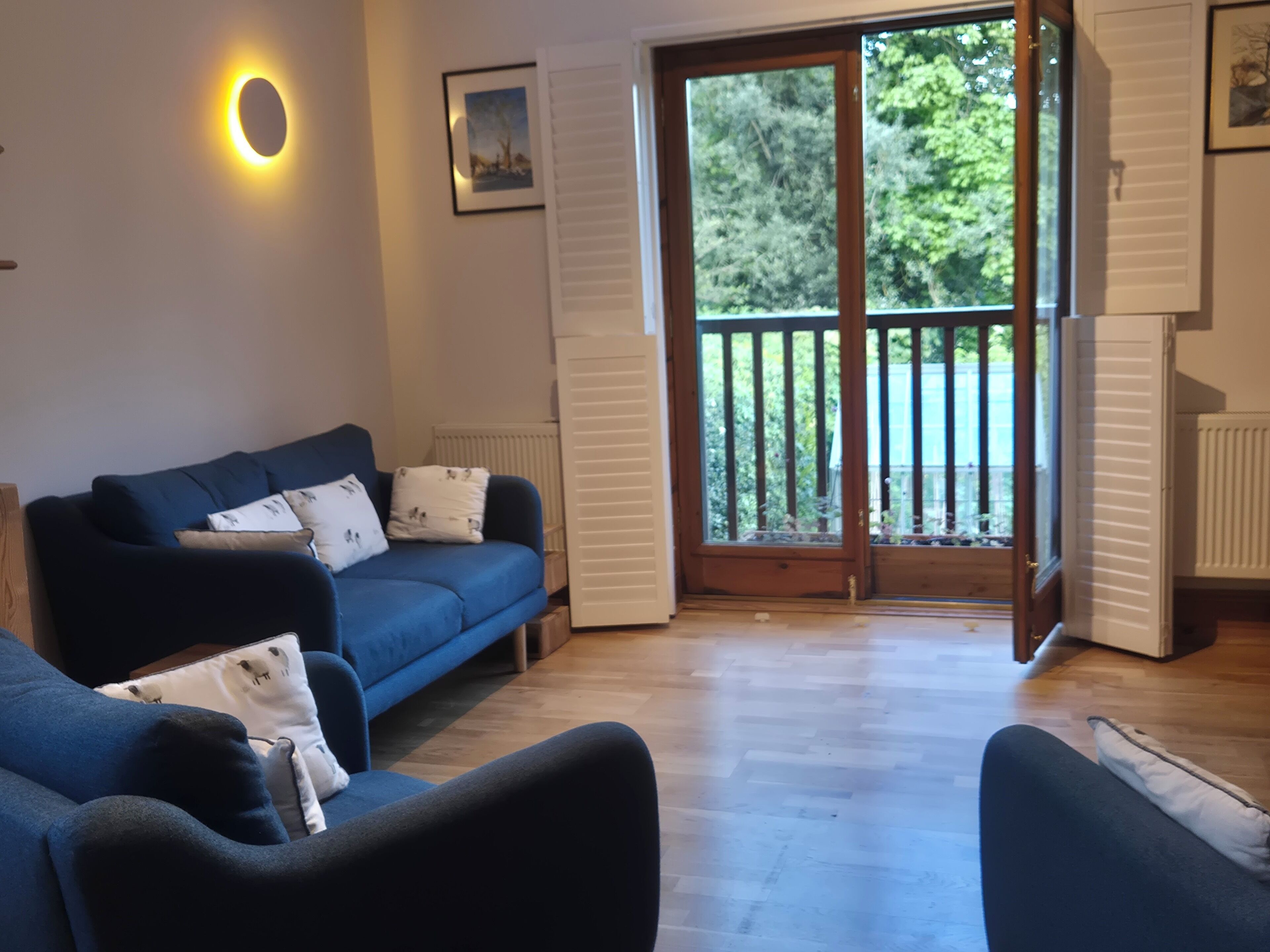 A living room with a blue sofa, a wooden floor, and a large sliding glass door leading to a balcony with a view of trees.