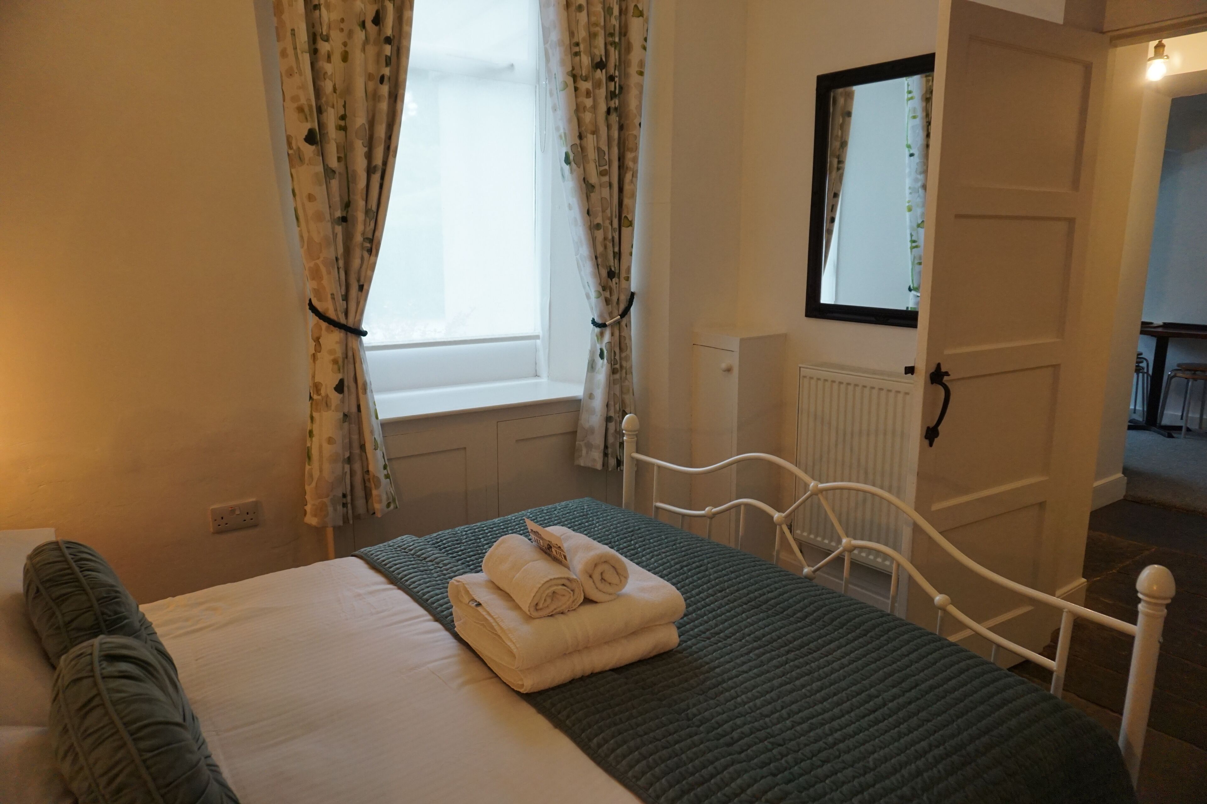 A neatly made bed with a green quilt, white linens, and rolled towels. There is a window with floral curtains, a white radiator, and a door leading to another room.