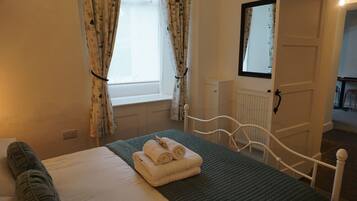 A neatly made bed with a green quilt, white linens, and rolled towels. There is a window with floral curtains, a white radiator, and a door leading to another room.