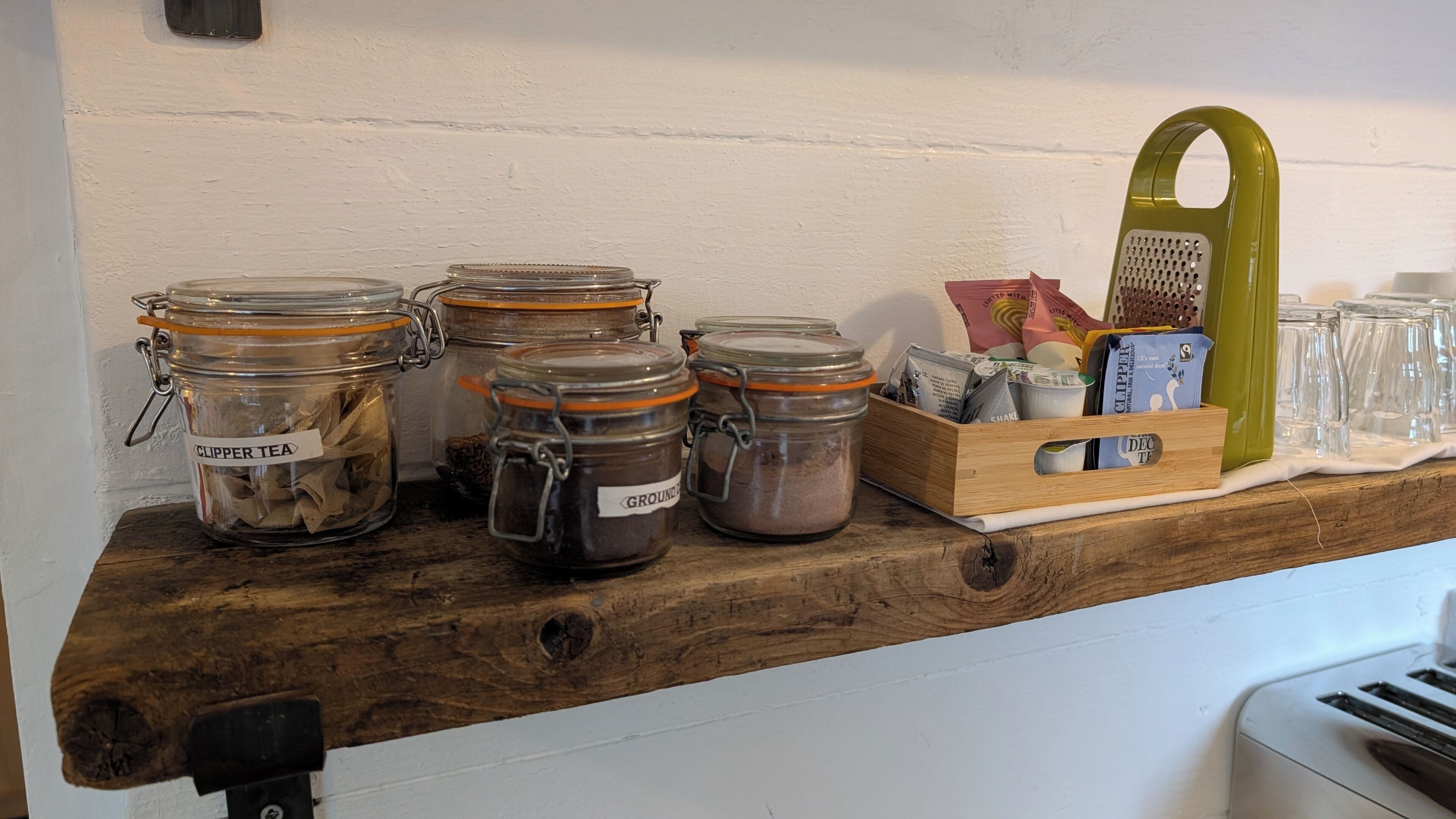 Una prateleira de fusta amb envasos de vidre etiquetats com 'GLIPPER TEA' i 'GROUND', una caixa de fusta amb bosses de te, i una tetera verda.