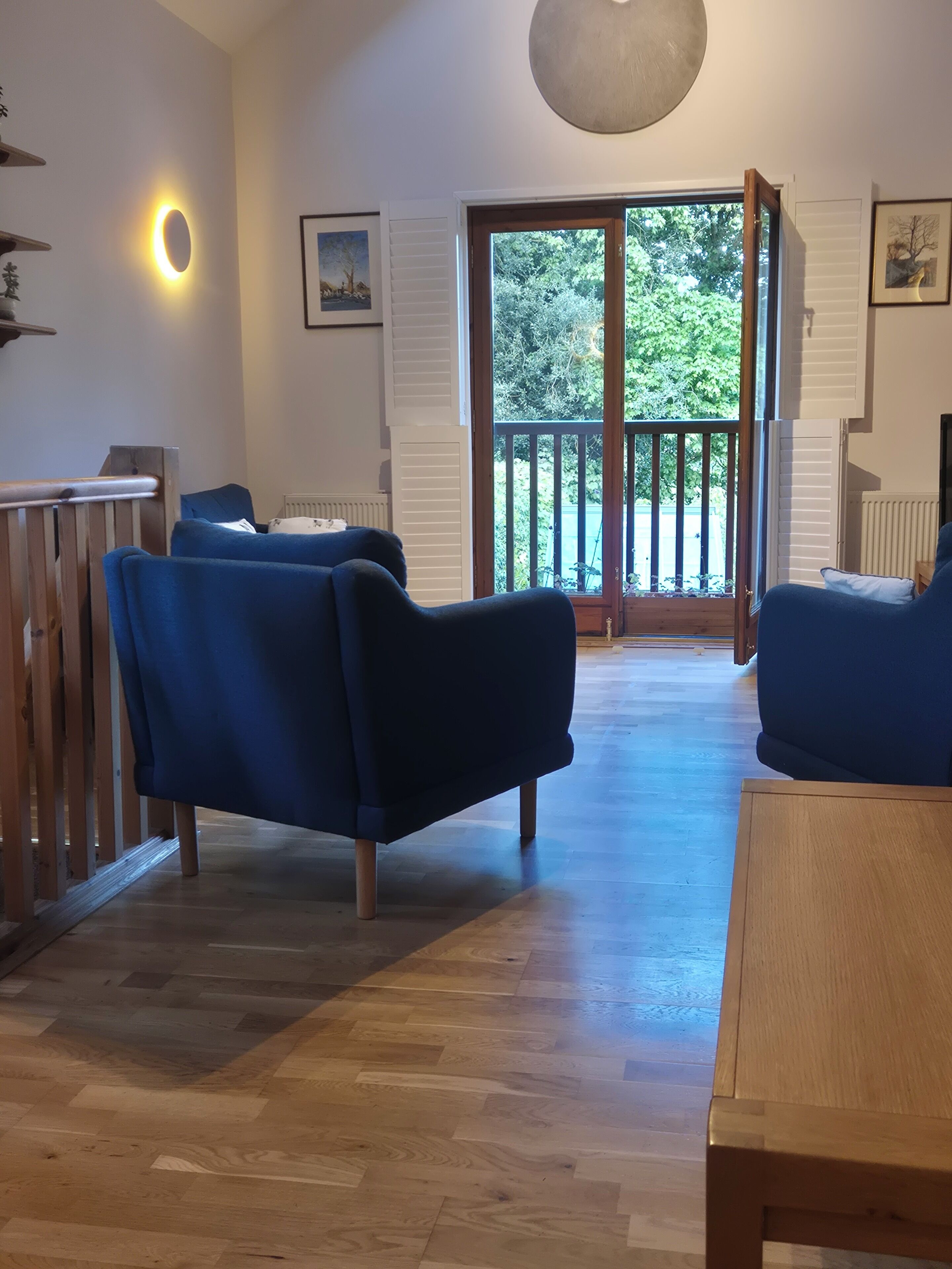 A room with a wooden floor, two blue sofas, a wooden table, and a balcony with a view of trees.